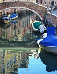 Fototapeta na wymiar Some motor boats are moored near a stone bridge on a Venetian canal. The boats and nearby buildings are reflected in the rippled water.
