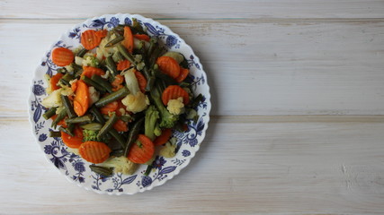steamed vegetables on a white plate with blue ornament on a white empty wooden surface top view