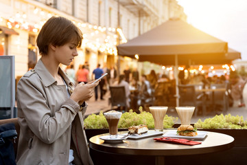 Millennial hipster teen girl blogger hold smart phone take food photo on phone mobile camera sit in city cafe at table. Young female vlogger shoot social media video story blog on smartphone outdoor. - obrazy, fototapety, plakaty