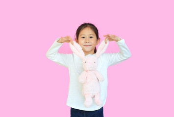 Adorable asian kid girl holding ears of pink rabbit doll isolated on pink background. Happy child on Easter