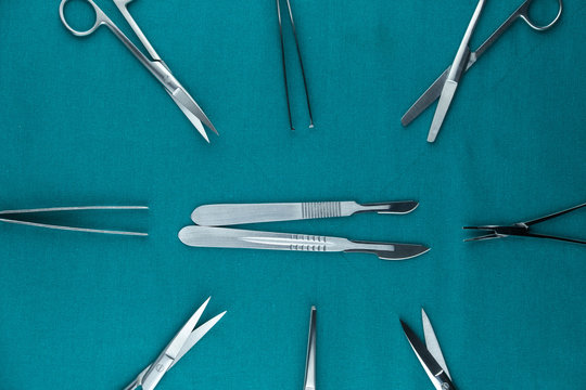 Top view of surgical instrument, stainless steel scalpel handle number 3 with blade number 10 and scalpel handle number 4 with blade number 24 and tweezers, scissors on surgical green drape fabric.