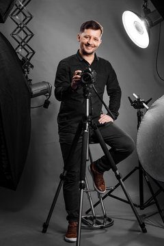 Happy man photographer with camera on a studio equipment background. Portrait of a smiling male model in black fashion clothes taking photos in the studio. Self portrait