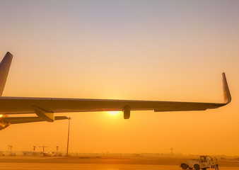Fototapeta na wymiar Closeup airplane wing with golden sunrise sky. Plane parked at airfield of airport. Morning flight. Summer travel concept. Pushback tractor support with airport staff at taxiways. Aviation business.