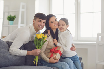 Happy mother's day. Father and baby daughter congratulates mom with flowers and a postcard in a room