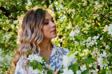 Naklejka premium Portrait of a beautiful blonde girl with green eyes and blossoming apple tree with white flower in the park in a spring time