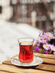 Tea in a glass of armudu on a saucer, located on a wooden table in the loggia. In the background is a bouquet of wildflowers. Bright sunny day. Close-up.