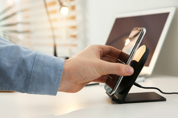 Man putting mobile phone onto wireless charger at white table, closeup. Modern workplace accessory