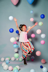 Fototapeta premium Happy asian kid girl in stylish striped maxi skirt has fun jumps skipping dances surrounded by colorful air balloons