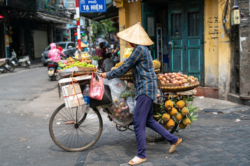 The street vendor with bike loaded of tropical fruits in old town street in Hanoi, old houses and...