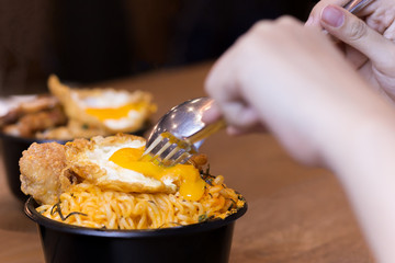 Hand holding spoon and fork cutting egg with noodle soup on wooden table.