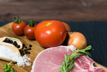 Raw pork chop meat on wood plate with vegetables, spices, salt and black pepper in dark black cloth table. Focus on tomato ingredient