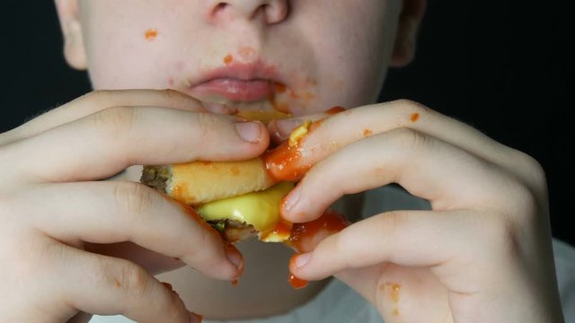The mouth of a teenager boy who eats a juicy burger a cheeseburger or hamburger with cheese, meat cutlet and pickles, dirty hands and face in ketchup. Unhealthy Fast Food and Street Food.