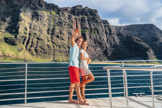 Hawaii cruise ship couple tourists taking selfie with mobile phone at Na Pali coast famous attraction in Kauai island, majestic dramatic landscape.