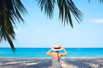 Kid in swimsuit and straw hat standing on the sea beach with palm tree