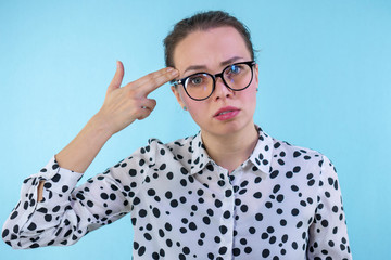 Woman with glasses shows a gesture of suicide on a blue background. Shooting and killing oneself pointing hand and fingers to head like gun.