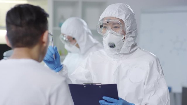 Tracking of unrecognizable female Asian doctor in disposable coveralls, protective mask and goggles talking to contagious male patient and writing down his symptoms, then checking his vision