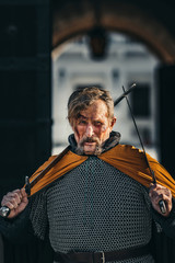 Portrait of a medieval senior warrior in armor after a battle with blood on his face. The warrior holds two swords in his hands