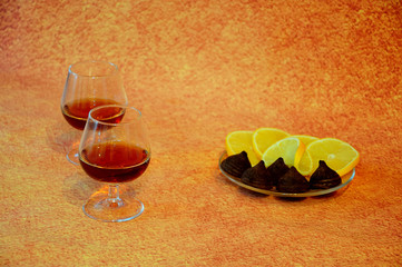 Two glass glasses with cognac and a plate with chocolate candies and cut lemon on a brown abstract...