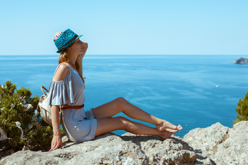 A young woman in a summer dress sits on the edge of a mountain or cliff with a beautiful sea view