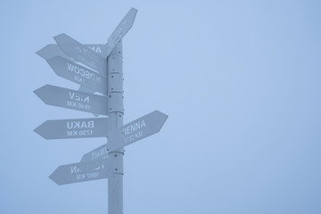 Gray direction sign pointing on capitals of countries on a gray foggy sky background. Tahtali mountain's peak, Turkey.