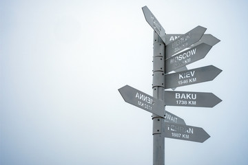 Gray direction sign pointing on capitals of countries on a gray foggy sky background. Tahtali mountain's peak, Turkey.