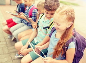 primary education, friendship, childhood, technology and people concept - group of happy elementary school students with backpacks sitting on bench and talking outdoors