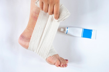 Woman is wrapping her leg with sprained ankle with elastic bandage isolated on white background. Twisted bandaged ankle and gel on white background.  Athlete runner is applying gel after  training acc