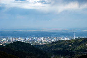 Fototapeta premium Beautiful view to Almaty city and foothills with storm clouds captured from mountains. Green almaty mountains foothills clouds in beautiful style on blue background.