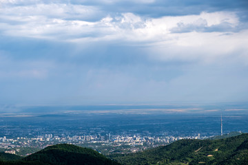 Fototapeta premium Beautiful view to Almaty city and foothills with storm clouds captured from mountains. Green almaty mountains foothills clouds in beautiful style on blue background.