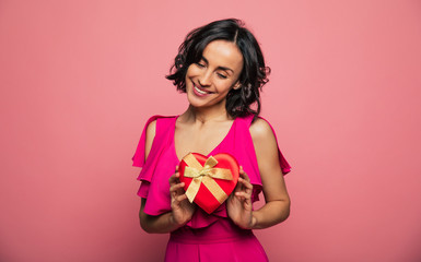 Cute gorgeous brunette woman is posing with a gift box in the shape of a heart in hands isolated on pink background.