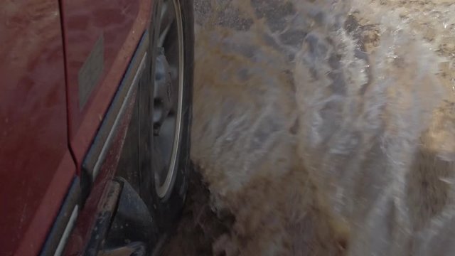 View of the tire on a red car as it drives through a muddy puddle in slow motion.
