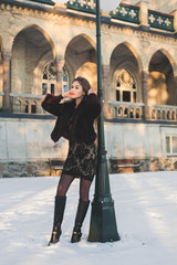 Fototapeta premium Fashion model with red lips under street light posing and watching a sun. Sunlight on the background. Snow around.