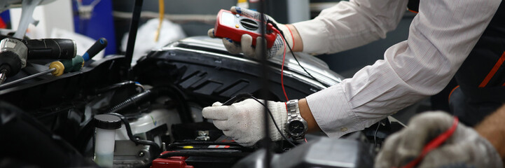 Close-up of male hands holding modern utility and checking voltage of automobile battery under hood of modern machine with accuracy. Automotive checkup concept