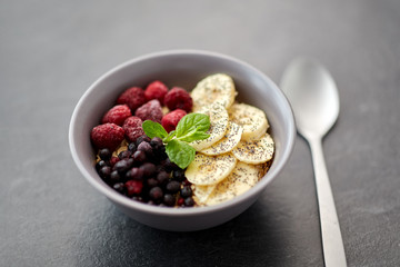 food and breakfast concept - oatmeal cereals in bowl with wild berries, banana and spoon on slate stone table