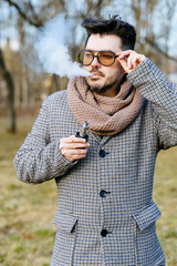 Confident young bearded male in sunglasses vape electronic cigarette. A young handsome bearded hipster man vaping e-cigarette outdoors in the park. Close up. - 328474397