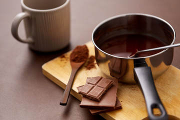 sweets, confectionery and culinary concept - pot with melted hot chocolate, cocoa powder in spoon, ceramic mug and wooden board on brown background