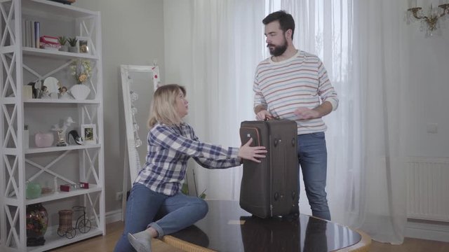 Proud Caucasian young man pushing wife's hands away from travel bag and turning back to woman. Confident husband leaving spouse, Divorce, conflict, breakup.