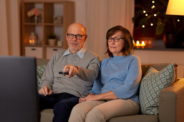 holidays, drinks and people concept - senior couple with remote control watching tv at home in evening