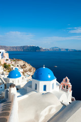 Fototapeta na wymiar Whitewashed Greek church featuring bright blue dome and pink bell tower overlooking a Mediterranean blue caldera view in Santorini, Greece