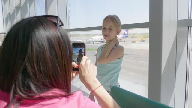 Young mother makes a photo of her daughter on the phone at airport while waiting for their flight. Traveling abroad with kids.