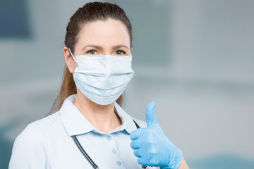 female doctor with medical face mask and medical gloves shows thumbs up 