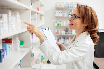 Smiling mid adult female pharmacist holding a clipboard and checking medication boxes on the...