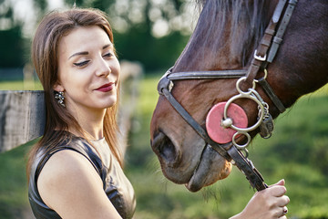 A young brunette woman in a black dress communicates with a dark brown horse on a sunny summer evening. Evening walk with a horse. Close-up.