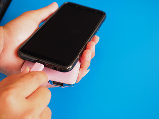  Close up woman's hand holding a smartphone is charging with a power bank on a blue background. Copy space. 