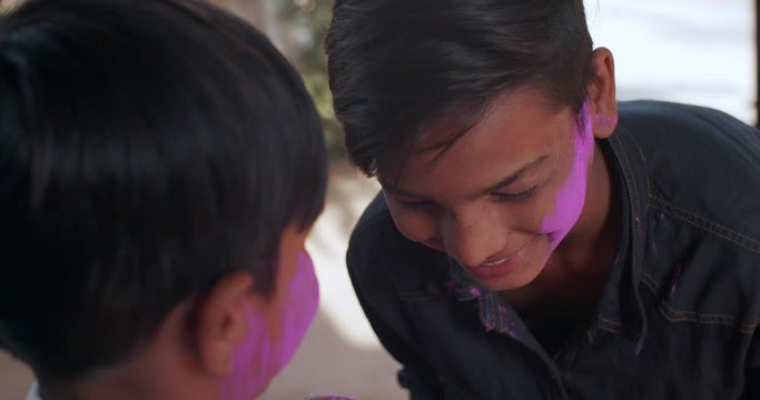 Two brothers, younger elder, playing Holi, traditional Hindu festival of colour in India by putting pink powdered color on each other's face cheeks and laughing and smiling, handheld slow-motion