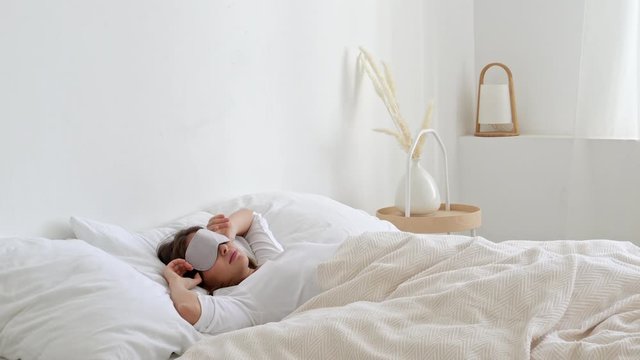 Pretty smiling young woman waking up, lying in bed in the moring, stretching arms, starting new day in minimalist white bedroom