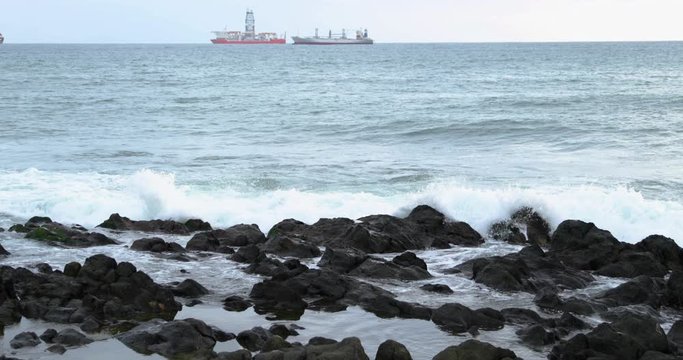 Waves crashing into a rocky shore full of large rocks and breaking water leaping to the sides with a pair of boats moored on the horizon at sea 4k slow motion capture at 60fps