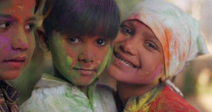 Close-up CU of sisters with younger brother hugging smiling excited looking straight at camera with colored faces in pink, green. orange, red celebrating Holi festival in India, handheld bokeh