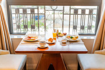 Fresh breakfast table next to window with bread, pastry, egg, fruit, juice, coffee cup