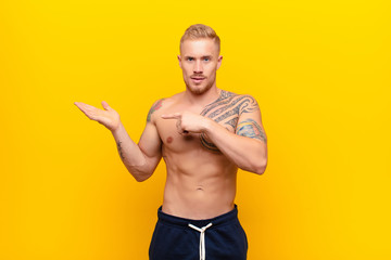 young strong blonde man smiling cheerfully and pointing to copy space on palm on the side, showing or advertising an object against yellow wall